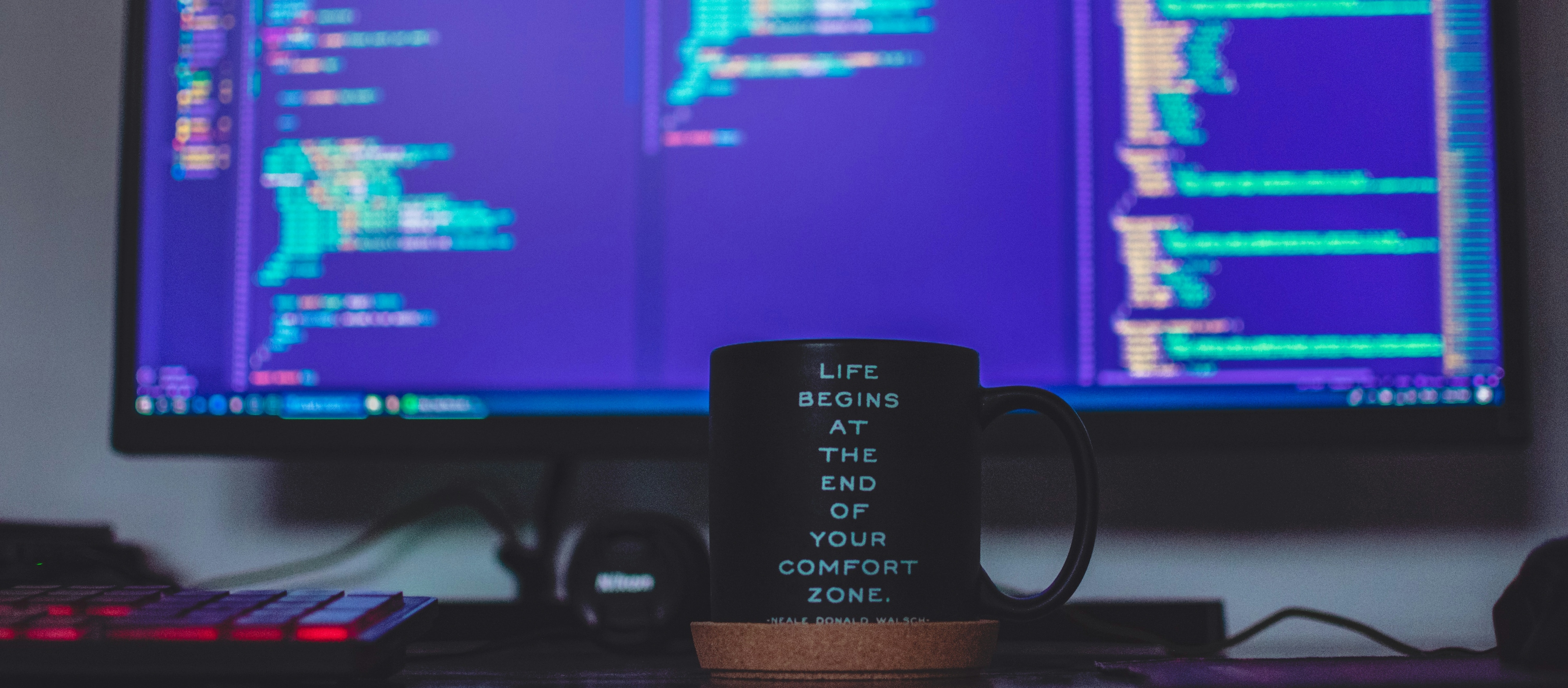 Picture of a remote working environment with a monitor with code and a mug that says 'Life begins at the end of your comfort zone'